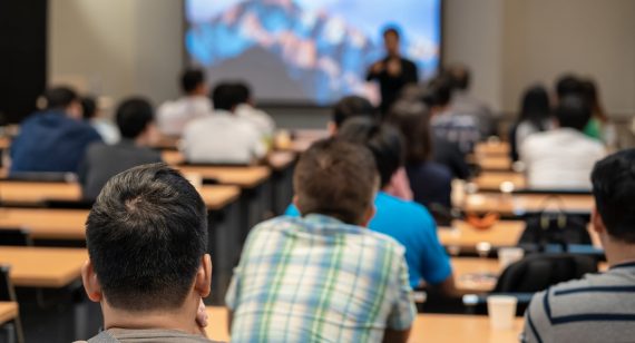 Speaker on the stage in front of the room with Rear view of Audience in put hand up acton for answer the question in the meeting or seminar meeting, business and education concept